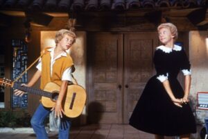 Hayley Mills portraying both twins, Susan and Sharon, in a lively scene from Disney’s 1961 film The Parent Trap. She plays guitar opposite her other character in a playful moment showcasing the film’s innovative split-screen effects.