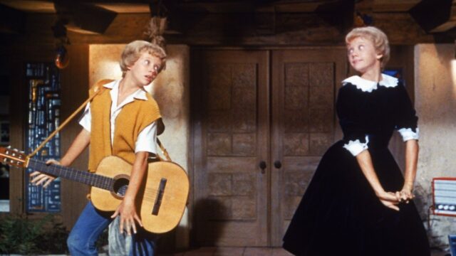Hayley Mills portraying both twins, Susan and Sharon, in a lively scene from Disney’s 1961 film The Parent Trap. She plays guitar opposite her other character in a playful moment showcasing the film’s innovative split-screen effects.