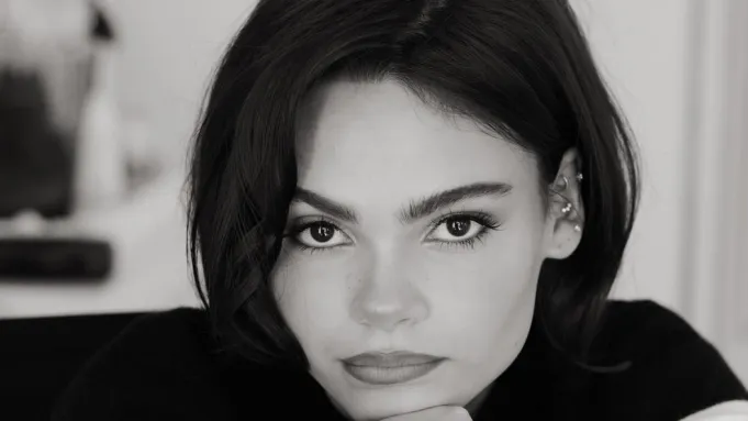 Black-and-white portrait of Ariana Greenblatt resting her chin on her hand, looking directly at the camera.