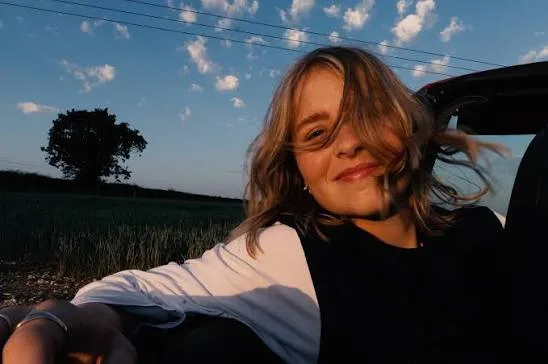Little Dog Star, the artist Izzy Steele, smiling while leaning out of a car window at sunset with an open field and a single tree behind her.