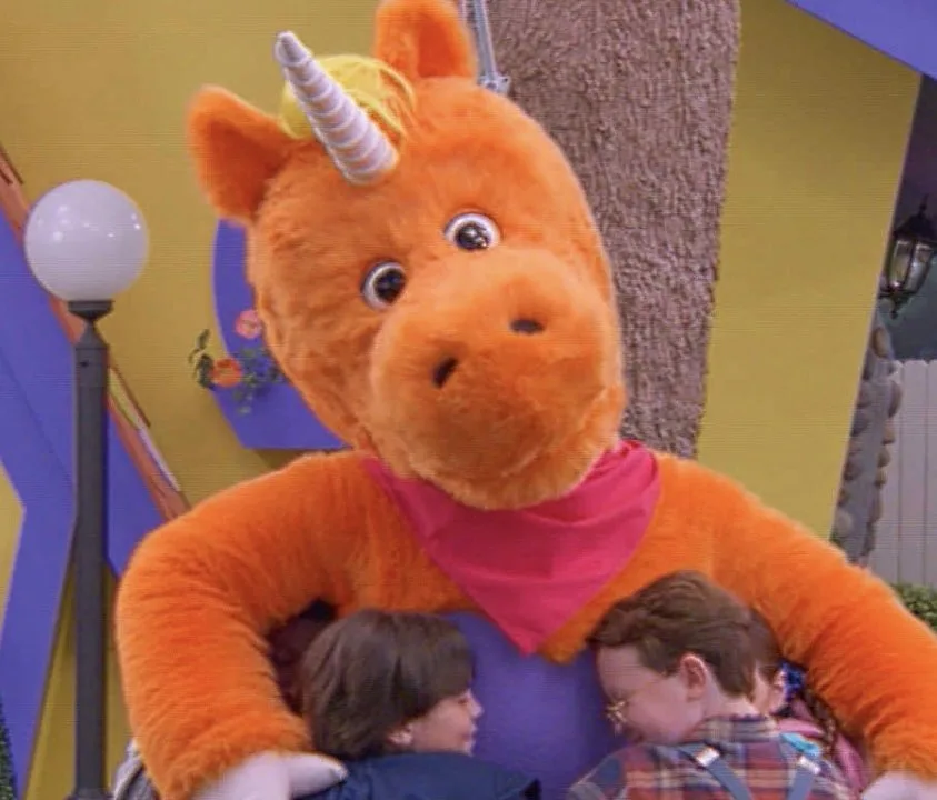 A large orange unicorn character wraps his arms around two children on a brightly colored children's television set.