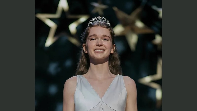 A smiling young woman wearing a silver tiara and a white dress stands in front of large glowing star decorations at a prom