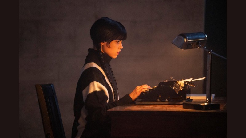 Jenna Ortega as Wednesday Addams typing on a vintage typewriter in a dark room