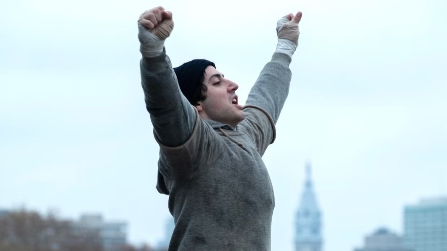 Anthony Ippolito as Sylvester Stallone with both arms raised, wearing gray sweats and a black beanie, Philadelphia skyline behind, in I PLAY ROCKY