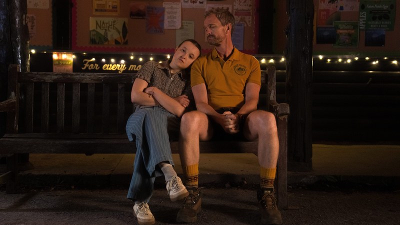 Bella Ramsey as Ivy leans on Neil Patrick Harris as Patrick on a camp bench at night in Sunny Dancer