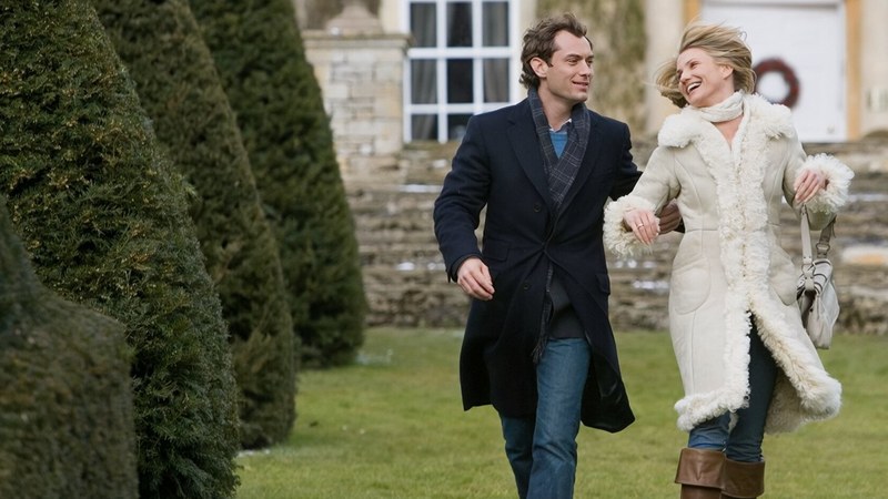 A smiling couple walks arm in arm through a garden outside a stone house, in a scene from The Holiday